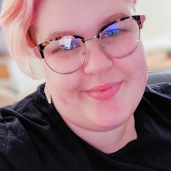 melissa looking at the camera with a smile, has bright pink hair, glasses and a black t-shirt